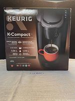 Front view of the Keurig K-Compact coffee maker packaging showing brand, model, key features and an image of the coffee maker with a red mug.