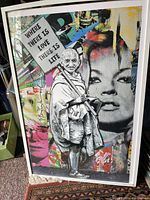 Full angled view of framed print poster leaning against wall on carpeted floor showing multicolored collage and white frame.