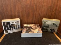Two unopened Blues CD box sets and one Blues book placed on a table with a wood background.