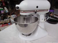 Front view of white KitchenAid Classic Ultra Power mixer with stainless steel bowl attached, placed on a white surface with various background items.