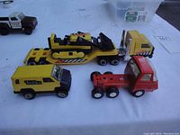 Four mini Tonka toy vehicles on white surface including a yellow van, a red truck, and yellow and black tractor-trailer with bulldozer.