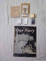 Overview of three Zippo lighters, a brown envelope with a military ribbon medal, small booklet 'Three Shall See', and 1939 'Our Navy' magazine.