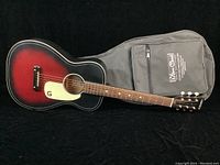 Full length photo of Gretsch Jim Dandy G9500 acoustic guitar with sunburst finish lying flat next to grey soft carry bag with logos.