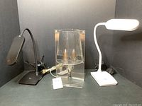 Photo showing the three lamps side by side: the vintage clear acrylic Kartell lamp in center flanked by the black and white flexible neck lamps.