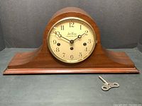 Front view of wooden mantle clock with brass bezel, cream face with Arabic numerals, and winding key placed beside it.