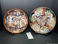 Two vintage Japanese Imari chargers displayed on stands, showing full front views of both plates with distinct colors and patterns.