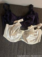 Two bras laid flat on a surface: one dark purple with lace details, the other cream colored.