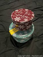 Two stacked round cookie tins with snowflake designs and winter holiday theme; red tin on top with 'Tis the Season' text and light blue tin underneath.