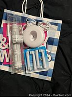 Electric toothbrush charger base, replacement brush heads, and clear protective cover grouped on a white and blue shopping bag on a black surface.