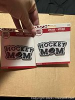Photo showing two individual 'Hockey Mom' iron-on patches in their original packaging, one with black lettering and one with red lettering.