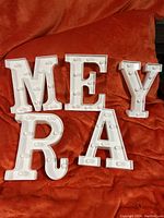 Five white light-up letters M, E, Y, R, A on an orange fabric surface. Letters have bulb sockets but are not lit.