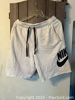 White Nike shorts hanging on a hanger showing front with black drawstring and large black Nike logo on left leg.