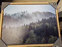 Front-facing photo of a framed oil painting showing a misty forest scene with dense trees and fog, in a wood frame with protective corner covers.