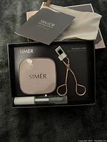 Top view of open SIMÉR eyelash kit showing the magnetic eyelash case, eyelash curler, and adhesive glue inside black packaging insert.
