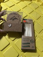 Photo shows unopened box of 50 natural wheat drinking straws alongside a new set of cork coasters in plastic packaging, placed on a yellow textured surface.