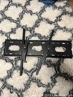 Black metal TV mount bracket with vertical mounting arms lying flat on a white and gray patterned rug.