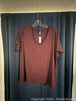 Dark red women's V-neck t-shirt hanging on a clear plastic hanger against a dark curtain background, tag attached to neckline indicating new condition