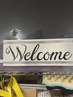Rectangular metal welcome sign with black cursive text and black border lines on white background, displayed on a shelf.
