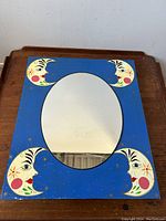 Front view of the rectangular mirror showing the blue wooden frame with four painted crescent moon faces at each corner and the oval mirror inset in the center.