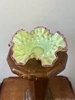 Side/top angle photo showing the crimped fiery vaseline glass bowl with cranberry rim on a wooden stand under natural light.