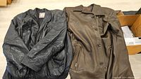 Two men's leather jackets laid flat on floor; left is black leather jacket, right is brown leather car coat.