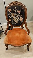 Full view of antique parlor chair showing wooden frame, embroidered floral backrest, and orange velvet seat.