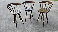 Three heavy wood swivel bar stools shown outdoors on pavement. Each stool has a contoured seat, spindle back with a cutout handle, and a metal ring footrest. Visible wear and scratches on wood.