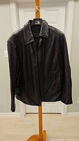 Full front view of the black leather jacket hanging on a wooden stand in front of white doors.