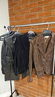 Three ladies jackets hanging on rack against brick wall: black leather car coat, black suede jacket, brown suede jacket