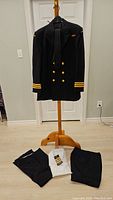 Navy wool double-breasted coat with gold buttons and sleeve rank stripes displayed on a hanger, three pairs of navy trousers and white shirt and navy tie below on floor.