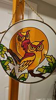 Close-up of stained glass window decoration showing two owls perched on a branch with detailed leaves and amber background, hanging by a chain.