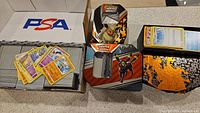 Large storage boxes filled with Pokémon trading cards with scattered cards placed on top including yellow-bordered cards, showing various Pokémon and moves.