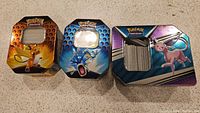 Three metal card tins featuring Pokémon characters Raichu, Gyarados, and Espeon, partially open showing traditional card slots inside.