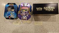 Three Pokemon items displayed: two tins with distinctive artwork and one black box labeled Shining Fates