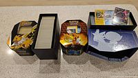 Three Pokémon card tins with most cards stored inside, some cards are displayed on top and in front showing variety and condition