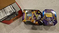 Side-by-side display of two metal Pokemon tins featuring Pokemon artwork alongside a box filled with digital code cards.