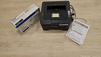 Brother printer with attached power cord, sticky note on top, sitting next to toner cartridge box and printed manual plastic wrapped.