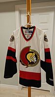 Front view of the Ottawa Senators white NHL jersey hanging on a hanger, showing the Senators logo and color details.