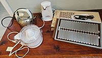 Four small kitchen appliances on wooden table: hot tray, electric juicer, can opener, electric kettle.
