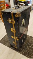 Side angled view of black metal steamer trunk with brass latches, metal reinforcements, and leather handle on top. Shows age wear and old stickers.
