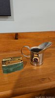 Photo showing the green-stone lighter and miniature metal jug with Royal Navy emblem side by side on wooden surface.