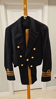 Photo of a black double-breasted navy uniform jacket with gold buttons and sleeve insignia on cuffs, hanging on a wooden stand in front of a white door.