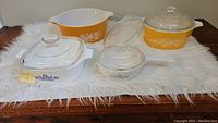 Sets of 5 Pyrex and Corning Ware glass bakeware pieces on table with white furry cloth underneath, showing yellow Butterfly Gold and white Cornflower Blue patterns with lids.