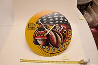 Front view of round Ed Hardy clock with bulldog design and white hands, next to a ruler for scale.