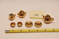 Three pieces of miniature china tea set in gold color displayed with measuring tape for scale, including a teapot, sugar bowl, and creamer.