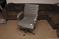 Gray swivel office chair shown from front angle on carpeted floor with brown sectional sofa in background.