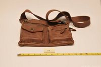 Front view of brown Roots leather shoulder bag with two front zippered pockets and adjustable strap. Measuring tape laid below the bag for size reference.