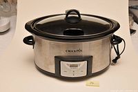 Front view of Crock-Pot slow cooker with stainless steel body, black side handles, digital control panel, and power cord attached, placed on a white surface with a sticky note labeled '26'.