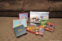 Photo showing all items in the lot placed on carpet near sofa: Hockey Night in Canada board game, Hangman, Knex set, Super Loops, Trivial Pursuit, Yahtzee, and 1000 piece puzzle