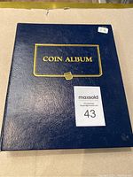 Front cover of dark blue vinyl coin album with gold “Coin Album” text, showing MaxSold lot sticker.
